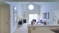 This single-floor space makes a strong architectural impression through its dramatically vaulted ceilings, which lend an airy, expansive quality to the main living area. A distinctive circular window positioned high on the front wall serves as a focal point, flooding the room with abundant natural light and adding a unique design element to the otherwise clean, white interior.

The living area is thoughtfully laid out, anchored by a fireplace set into a recessed niche along one wall — a practical and visually grounding feature. Sliding glass doors on the opposite wall open to an outdoor area, further enhancing the sense of openness while providing exterior views and additional light. The neutral carpet throughout softens the space and complements the all-white walls and trim.

A partial counter or peninsula transitions toward what appears to be a kitchen or dining area, suggesting an open-concept floor plan that encourages easy flow between spaces. Framed artwork lines one wall, adding dimension without disrupting the minimalist aesthetic. Overall, the space feels bright, clean, and well-proportioned, with architectural details that elevate it beyond the ordinary.