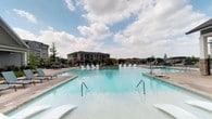 This outdoor amenity space centers around a generously sized resort-style swimming pool, offering a leisure-focused environment designed for relaxation and recreation. The pool features sparkling, light-blue water with a smooth, clean finish that reflects the open sky above. Entry points with chrome handrails are visible at multiple access areas, making the pool accessible and inviting.

Surrounding the water, a spacious pool deck is lined with a generous arrangement of lounge chairs and chaise lounges, providing ample space for sunbathing and unwinding. The layout allows for comfortable spacing between seating areas, contributing to a resort-like atmosphere. Stone or textured accent details are visible along portions of the pool's edge, adding a refined, decorative touch to the hardscape design.

The backdrop features multi-story residential buildings with covered balconies, mature trees, and open landscaping that frame the space and provide visual depth. The setting benefits from an expansive open sky, allowing abundant natural light to illuminate the area throughout the day. The overall ambiance is bright, airy, and well-appointed, reflecting a community-focused outdoor retreat.