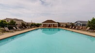 Located at 56 Bent Branch Loop in Clayton, NC, this single-level community amenity space is thoughtfully designed for outdoor living and social gatherings. At its centerpiece is a generously sized swimming pool with a dedicated pool house, surrounded by a wide concrete deck lined with lounge chairs and enclosed by a black wrought-iron fence.

Adjacent to the pool is a well-appointed covered pavilion that serves as the heart of the outdoor entertainment area. The pavilion features elegant white columns with stone bases and soaring arched ceilings finished in warm wood tongue-and-groove paneling. Ceiling fans are installed throughout to ensure comfort during warmer months. The space is divided into distinct functional zones: a lounge area with cushioned seating arranged around a coffee table, a dining area with a decorative cast-iron table and chairs, and an outdoor fireplace wall clad in natural stone with a rustic wood mantel and a mounted flat-screen television above it.

French doors connect the covered pavilion to an interior structure, offering seamless indoor-outdoor access. The open-air design provides unobstructed views of the pool and surrounding community landscape, creating a welcoming and versatile space ideal for both casual relaxation and organized community events.