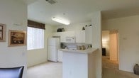 This single-story space features an open-concept kitchen that serves as a central hub of the layout. The kitchen is outfitted with white cabinetry that extends to the upper walls, providing generous storage capacity. Upper cabinets reach toward the ceiling, offering additional shelving space currently used for decorative plants and accents. White appliances include a full-size refrigerator, electric range, and built-in microwave, all seamlessly integrated into the cabinetry. A peninsula-style breakfast bar extends from the counter, creating a natural divider between the kitchen and the adjacent living area while offering casual seating or prep space. The countertops are light-toned and clean in appearance. A large window fitted with a bamboo roller shade allows natural light to filter into the kitchen area, brightening the space. Flooring in the kitchen appears to be a light-toned hard surface, transitioning to carpet in the adjoining living area. The overall color palette is neutral, featuring soft whites and beige tones throughout. A hallway visible beyond the living area suggests additional rooms within the unit. Overhead fluorescent lighting provides consistent illumination throughout the kitchen.
