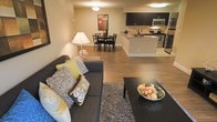 This single-floor residence offers a thoughtfully designed open-concept layout that integrates the living, dining, and kitchen areas into one cohesive and functional space. The living room anchors the main area with warm hardwood-style flooring, a plush sectional sofa accented with decorative pillows, and a sleek coffee table — all centered around a textured area rug that adds depth and comfort underfoot. Warm ambient lighting from table lamps and ceiling fixtures creates an inviting atmosphere throughout.

Flowing seamlessly from the living area, the dining space accommodates a four-person table and chairs, providing an intimate setting for meals. The open sightlines between areas enhance the sense of spaciousness and encourage easy interaction across the floor plan.

The kitchen is a standout feature, boasting dark espresso cabinetry that contrasts elegantly with light countertops. A peninsula with bar-height seating serves as both a prep station and casual dining spot. Stainless steel appliances — including a microwave, range, and refrigerator — complete the modern aesthetic. Neutral warm-toned walls tie the entire space together, lending a cohesive, welcoming ambiance throughout this well-balanced and stylishly appointed home.
