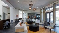 This sophisticated single-floor space functions as a contemporary clubhouse or community lounge, thoughtfully designed to blend comfort with modern elegance. The open-concept layout creates a generous sense of space, anchored by soaring ceilings that amplify the airy ambiance throughout.

The main lounge area is furnished with plush white sofas and accent chairs arranged around bold, dark circular coffee tables, all resting on a vibrant patterned area rug in warm amber and gold tones. This color palette adds a striking contrast against the neutral grey tile flooring and crisp white walls.

Overhead, a trio of sculptural wood-slat pendant lights serve as a dramatic focal point, casting a warm, layered glow that complements the recessed lighting throughout. A dedicated coffee and refreshment bar lines the back wall, appointed with countertop seating and stocked with beverage service amenities, making it ideal for casual gatherings.

Behind the bar, a textured stone accent wall frames a mounted television, adding depth and a refined, upscale character to the space. Tall windows dressed in floor-length curtains allow natural light to filter in while maintaining an intimate atmosphere. Additional seating areas are visible in adjacent zones, offering versatile spaces for socializing or quiet relaxation.
