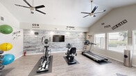 Located in Glendale, Arizona, this dedicated fitness center offers a comprehensive workout environment within a compact, efficiently designed single-room layout. The space is defined by striking vaulted ceilings that create an open, airy atmosphere, complemented by two modern ceiling fans that promote airflow throughout the room. Light wood-style flooring runs underfoot, lending a clean and contemporary aesthetic.

The cardio zone is anchored by a treadmill, an elliptical trainer, and a stationary cycle, each positioned on protective floor mats. A flat-screen television is wall-mounted on a textured, whitewashed brick accent wall — a standout architectural feature that adds visual character and serves as a motivating focal point. Motivational phrases and fitness slogans are displayed across the walls, reinforcing an energetic workout environment.

Strength training is well-supported with a rack of hex dumbbells in graduated weights, a set of exercise balls in multiple sizes mounted on a vertical storage rack, and a multi-function cable resistance tower equipped with various attachments. An adjustable weight bench provides additional versatility for strength exercises.

Natural light enters through multiple windows fitted with blinds, offering views of the surrounding outdoor greenery. The space is thoughtfully organized to maximize usability, making it a fully functional community fitness facility.