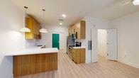 This single-floor residence showcases a thoughtfully designed, modern interior with a cohesive aesthetic throughout. The open-concept kitchen serves as the heart of the space, featuring warm-toned wood cabinetry, crisp white quartz countertops, and a classic white subway tile backsplash. A peninsula island provides additional prep space and casual seating. Stainless steel appliances — including a refrigerator, over-the-range microwave, and electric range — add a polished, functional touch. Pendant lights hang above the kitchen, complementing the recessed lighting overhead. A bold teal accent door adds a pop of color and character. Light-toned hardwood-style flooring runs seamlessly throughout the entire space, creating a sense of continuity and warmth. The bedroom is bright and airy, benefiting from a large window that invites natural light and features a ceiling fan for added comfort. The bathroom maintains the clean, minimalist theme with white cabinetry, a vanity mirror, and continued light flooring. The overall ambiance is fresh and contemporary, with a neutral white palette offset by natural wood tones, making the space feel inviting, well-lit, and move-in ready.