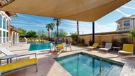 Located at 11410 Edgemere Blvd in El Paso, Texas, this single-level outdoor amenity space offers a well-appointed resort-style pool area designed for relaxation and recreation under the West Texas sun. The centerpiece is a generously sized swimming pool with crystal-clear blue water, complemented by a dedicated spa and a striking stone water feature that adds both visual interest and a soothing ambient sound. The pool deck is finished in a warm concrete surround and features multiple tanning ledges and in-water platforms, providing ample space for sunbathing. Bright yellow lounge chairs and seating arrangements are distributed throughout the deck, offering both open-air and shaded options. A large modern shade sail stretches over a portion of the deck, providing a comfortable covered retreat from direct sunlight. The space is enclosed by a privacy wall and bordered by mature, tall palm trees and desert-adapted plantings, creating a lush, tropical atmosphere. Adjacent to the pool, a covered outdoor pavilion with classic arched colonnades, red-tile roofing, and large windows serves as an architectural focal point with a distinct Mediterranean flair. The overall ambiance is vibrant yet relaxed, making this outdoor space a standout communal feature for the surrounding residential community.