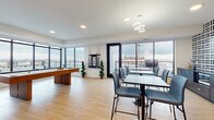 This single-floor amenity lounge is designed as a versatile social and recreation space, combining thoughtful interior design with expansive city views. Light hardwood flooring runs throughout, complementing the clean white ceilings equipped with recessed lighting and a contemporary circular chandelier above the dining area.

The space is divided into distinct functional zones. A game area anchors one side, featuring a full-size pool table and a shuffleboard table alongside a wall-mounted cue rack. Adjacent to the game zone, a cozy lounge nook is defined by a bold navy accent wall, textured bench seating, and small café-style tables with rounded accent chairs — an ideal spot for casual conversation.

The dining area offers high-top tables with sleek grey upholstered chairs, positioned directly in front of floor-to-ceiling windows that frame sweeping urban skyline views. A kitchenette and service area along one wall is accented by bold geometric patterned tile and navy cabinetry with brass hardware, paired with open display shelving styled with plants, books, and decorative objects.

Large sliding glass doors provide direct access to an outdoor terrace, filling the interior with natural light and further enhancing the open, airy ambiance throughout.