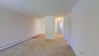 This single-floor space is characterized by its clean, neutral aesthetic and functional layout. The main living area features wall-to-wall beige carpeting that extends throughout, creating a cohesive and understated foundation. Crisp white walls reflect available light, keeping the space feeling open and airy despite its modest proportions.

A central partial wall divides the main living area from the connecting hallway, which leads toward additional rooms at the rear of the unit. The hallway is well-lit with ceiling-mounted fixtures, and a window visible at the far end introduces natural light deep into the interior. Along the perimeter of the main living area, a baseboard heating unit provides climate control.

To the right, a bi-fold door suggests a closet, offering practical built-in storage. The overall layout appears straightforward and efficient, with a clear circulation path connecting the living space to the rest of the unit. The uniformity of the flooring, wall color, and trim throughout gives the space a cohesive, move-in-ready appearance suitable for a variety of furnishing arrangements.