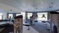 Located at 1600 Club Drive in Woodbury, NJ, this fitness center is a well-appointed, single-floor facility designed to support a well-rounded workout experience. The space is finished with a clean, contemporary aesthetic, featuring neutral gray walls, white ceilings, and durable flooring that transitions between zones within the room. Recessed lighting throughout the ceiling, complemented by a ceiling fan, ensures the space remains well-lit and comfortably ventilated during workouts. An abundance of natural light pours in through multiple large windows that line the walls, offering views of the surrounding greenery and creating an open, energizing atmosphere. The fitness floor is equipped with a variety of cardio machines, including multiple treadmills positioned along the windowed walls, as well as a stationary bike. Strength training is supported by a dedicated weight and resistance machine, accompanied by a workout bench for versatile exercise options. A full-length mirror along one wall enhances the sense of space and allows users to monitor their form. The layout is thoughtfully organized to provide ample room for movement between equipment, making this fitness center a practical and inviting amenity.