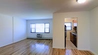 This single-floor space offers a clean, functional layout with a welcoming open-concept design that connects the living area to the kitchen. The living space is characterized by smooth hardwood flooring that reflects natural light pouring in through a wide multi-pane window, creating a bright and airy atmosphere. White walls throughout contribute to the sense of openness, while a baseboard heating system and a wall-mounted air conditioning unit ensure year-round comfort. A partial dividing wall separates the living area from the kitchen, maintaining an open feel while providing a sense of definition between spaces. The kitchen is well-appointed with warm wood cabinetry, a double sink, and black appliances including a refrigerator and dishwasher. Tile flooring in the kitchen offers a practical contrast to the hardwood in the living area. A secondary window in the kitchen brings in additional natural light, keeping the cooking space bright and pleasant. The overall ambiance is straightforward and functional, with neutral tones and practical finishes that offer a versatile canvas for personalization. The space is well-suited for everyday living, balancing comfort and utility across its single level.