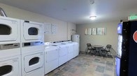 This single-floor communal laundry facility is designed for practicality and ease of use. The room features a row of full-sized front-loading washing machines paired with stackable dryers, providing ample capacity to accommodate multiple users at once. Informational notices are posted on the wall above the machines, ensuring guests and residents can easily reference operating instructions and guidelines. The flooring is finished in a durable, multi-toned stone tile that adds a grounded, clean aesthetic to the utilitarian space. A small seating area in the corner, furnished with a round table and folding chairs, offers a comfortable spot to wait while laundry cycles run. A colorful multi-panel artwork on the wall adds a touch of warmth and character to the otherwise functional environment. The room is well-lit with ceiling-mounted lighting, contributing to a bright and welcoming atmosphere. Overall, this laundry facility is thoughtfully arranged to balance efficiency and comfort, making routine laundry tasks straightforward and convenient.