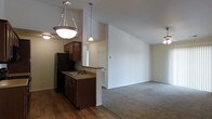 This single-floor residence offers an open, airy layout that connects the kitchen and living areas in a cohesive, functional design. The kitchen is outfitted with rich, dark wood cabinetry that provides ample storage, a double-basin sink, and a full-size refrigerator. A breakfast bar peninsula separates the kitchen from the rest of the space, offering a convenient spot for casual dining or food preparation. Pendant lighting hangs above the kitchen area, adding warmth and focused illumination, while a matching ceiling-mounted fixture reinforces the cohesive lighting scheme. The flooring transitions from smooth hardwood-style planks in the kitchen to plush carpeting in the living area, clearly delineating the two zones while maintaining an open feel. Vaulted ceilings rise dramatically overhead, enhancing the sense of spaciousness throughout. The living area is generously proportioned, offering flexible space for a variety of furniture arrangements. Neutral wall tones throughout create a clean, versatile backdrop that complements a range of interior styles. A ceiling fan in the living area adds both comfort and functionality. Overall, the space balances practicality with an inviting, open ambiance.