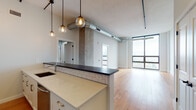 This urban loft showcases a compelling blend of industrial aesthetics and modern refinement across a single, open-concept floor. Warm hardwood flooring flows throughout, complementing the exposed concrete columns, visible ductwork, and black pipe accents that define the space's industrial character. Soaring ceilings amplify the sense of openness in the main living area, which is anchored by expansive floor-to-ceiling windows that frame sweeping city skyline views and flood the interior with natural light.

The kitchen is a standout feature, outfitted with white shaker cabinetry, subway tile backsplash, sleek quartz countertops, and a dark-topped peninsula that serves as both a prep surface and a casual dining bar. A suite of stainless steel appliances — including a gas range, built-in microwave, dishwasher, and refrigerator — rounds out the functional design. Globe pendant lights suspended at varying heights add warmth and a contemporary industrial touch.

The bedroom is generously proportioned, benefiting from its own set of large windows that offer elevated city views. The bathroom is finished with marble-look tile on both the floors and tub surround, paired with a dark-stained vanity, dark stone countertop, and a wall-mounted sconce — creating a clean, spa-inspired atmosphere. Together, these spaces deliver a cohesive, thoughtfully designed urban living environment.