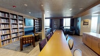 This single-floor multipurpose space is thoughtfully designed to accommodate both formal meetings and relaxed gatherings. At its center sits a long, warm-toned wooden conference table flanked by cushioned leather chairs, making it well-suited for group discussions, presentations, or collaborative work sessions. A textured ceiling with recessed lighting provides even illumination throughout the room, complementing the natural light that streams in through large windows dressed with dark draping curtains. The windows offer views of surrounding greenery, adding a calm, natural backdrop to the interior. Along one wall, floor-to-ceiling built-in bookshelves are filled with volumes, paired with a large wall-mounted display screen — an ideal setup for media presentations or research. Adjacent to the main conference area, a comfortable lounge zone features upholstered sofas and armchairs arranged around a side table, offering a more informal setting for conversation or relaxation. Additional seating clusters near the windows further enhance the room's flexibility. The flooring transitions between carpet in the lounge area and hardwood-style surfaces near the perimeter, adding visual distinction between zones. Framed artwork adorns the walls, contributing to a polished and professional yet welcoming ambiance throughout.