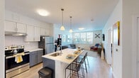 Located at 10 Patriot Parkway in South Weymouth, MA, this well-designed one-bedroom, one-bathroom residence offers a thoughtfully laid-out single floor of comfortable living space. The open-concept kitchen is a standout feature, showcasing two-tone cabinetry with crisp white upper cabinets and bold blue-gray lower cabinets, stainless steel appliances, a subway tile backsplash, and a large center island with a white quartz countertop and built-in sink — ideal for both meal preparation and casual dining. Light wood-style flooring flows throughout the main living areas, enhancing the bright, airy ambiance. The living room is generously proportioned, featuring large windows that invite abundant natural light and views of the surrounding neighborhood. The bedroom offers a cozy, well-sized retreat with plush carpeting, a window with outdoor views, and a dedicated closet for storage. The full bathroom is finished with gray subway tile in the tub/shower surround, tile flooring, a gray vanity with white countertop, and clean, modern fixtures. Throughout the residence, neutral white walls and contemporary finishes create a clean, modern aesthetic that is both inviting and versatile.