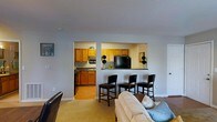 This single-floor residence offers a practical and welcoming layout designed for comfortable everyday living. The living area is open and inviting, featuring neutral-toned carpet and light gray walls that create a clean, airy atmosphere. A large sofa anchors the space, which flows seamlessly toward the kitchen area.

The kitchen is defined by warm honey-toned wood cabinetry paired with dark countertops, providing ample storage and workspace. A standout feature is the pass-through window that opens directly from the kitchen into the living area, enhancing the sense of connectivity between the two spaces. A built-in breakfast bar lines the pass-through, complemented by a set of dark bar stools — ideal for casual dining or entertaining.

The kitchen is well-equipped, featuring black appliances including a refrigerator and range, along with a sink positioned beneath upper cabinetry. Decorative touches such as small topiaries add a homey charm. A door off the kitchen provides convenient access to adjacent areas of the home.

Overall, the space strikes a balance between functionality and comfort, with a cohesive neutral color palette, thoughtful layout, and a warm, lived-in ambiance that makes it feel immediately welcoming.