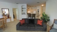 This single-floor residence offers an open-concept layout that connects the living, dining, and kitchen areas into one cohesive and functional space. The living area is carpeted and comfortably sized, anchored by a plush charcoal sofa accented with warm rust-colored throw pillows, creating an inviting atmosphere. A distinctive arched built-in shelving niche adds architectural character to the main living space, providing a decorative focal point ideal for displaying personal items.

The kitchen is situated just beyond the living area and features light wood cabinetry, a white refrigerator, and a gas range, offering a clean and practical cooking environment. Track lighting illuminates the kitchen, enhancing visibility and adding a modern touch. A raised breakfast bar with tall bar seating provides a casual dining option and serves as a natural transition between the kitchen and living spaces.

The open flow of the layout ensures easy movement throughout the unit, while interior doors visible from the main area suggest additional private rooms beyond. The overall ambiance is warm and approachable, with neutral wall tones and light-colored finishes throughout that contribute to a bright, airy feel. This well-appointed space balances comfort and functionality in a compact, efficient single-level design.