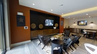 This single-floor space showcases a bold, contemporary design anchored by a striking media wall finished in deep black, set against warm terracotta-orange walls that create a vibrant, energetic atmosphere. The focal point of the lounge area is a wall-mounted flat-screen television framed by decorative woven circular wall accents and a low-profile wooden credenza below, providing both style and storage. A curated seating arrangement of dark leather mid-century-style chairs surrounds a geometric patterned area rug and a round black accent table, creating an inviting social setting ideal for gathering or relaxing. Recessed ceiling lighting, complemented by warm accent lighting along the upper wall transitions, ensures well-balanced illumination throughout. Light gray plank-style tile flooring runs throughout, adding a clean, modern contrast to the rich wall tones. In the background, a dedicated wet bar or kitchenette area is visible, complete with pendant lighting and cabinetry, offering added functionality. The overall ambiance is sophisticated and social, blending residential warmth with a modern hospitality aesthetic.