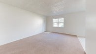 This single-story, two-bedroom unit offers a straightforward and functional layout designed for comfortable everyday living. The living area is generously sized, featuring neutral-toned wall-to-wall carpeting and a textured ceiling that adds subtle dimension to the space. A window allows natural light to filter in, brightening the open floor plan. A wall-mounted electrical panel is conveniently located within the main living space.

The galley-style kitchen runs along a narrow corridor, with cream-colored cabinetry lining both sides and wood-trimmed accents providing a warm contrast. It is equipped with a double-basin sink, an electric range, and an overhead range hood, along with ample upper and lower cabinet storage. The kitchen features tile-pattern vinyl flooring, distinct from the carpeted areas elsewhere in the unit.

The bedrooms are carpeted and maintain the clean, white-walled aesthetic found throughout the unit. Each room receives natural light from windows with outdoor views. At least one bedroom features a dedicated closet with a built-in rod for hanging storage. The bathroom includes a towel bar and a brushed nickel door handle, maintaining a simple and clean finish. Overall, the unit presents a bright, neutral, and move-in-ready environment.