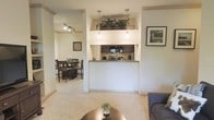 This single-story space showcases an open-concept design that seamlessly connects the living, dining, and kitchen areas. The kitchen is defined by a half-wall pass-through counter with a dark granite or stone surface, creating a natural boundary while maintaining visual openness. Two pendant lights hang above the counter, providing focused task lighting, while a decorative plant display atop the pass-through adds a touch of greenery and visual interest.

The living area features plush, light-toned carpeting and is furnished with a sofa and a round metallic coffee table, creating a relaxed and welcoming atmosphere. Framed artwork on the walls contributes to the curated feel of the space.

Adjacent to the kitchen, the dining area is accessible through a columned opening and is outfitted with a dark wood dining table and matching chairs. Track lighting overhead ensures the dining space is well-lit. The consistent use of soft white and neutral tones throughout the unit enhances the sense of openness and light, giving the overall space a clean, cohesive, and inviting character.