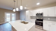 This one-bedroom, one-bathroom residence at The Bentley in North Little Rock, Arkansas offers a thoughtfully designed single-story layout with contemporary finishes throughout. The open-concept living and kitchen area is anchored by a large central island featuring a quartz countertop, double undermount sink, and stylish pendant lighting above. The kitchen is well-appointed with an abundance of white shaker-style cabinetry, a gray tile backsplash, and a full suite of stainless steel appliances, including a refrigerator, range, and over-the-range microwave. Light wood-tone flooring extends throughout the living areas, complementing the neutral wall tones and creating a clean, cohesive aesthetic. Recessed lighting enhances the bright, airy atmosphere of the main living space. The bedroom features large windows fitted with blinds, allowing ample natural light while maintaining privacy and offering views of the surrounding area. The bathroom is well-equipped with a wide white vanity, generous countertop space, a large mirror, and a tub-shower combination, all finished with consistent modern hardware. The overall design reflects a modern sensibility, with clean lines and a neutral palette that provides a versatile, move-in-ready environment.