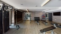 This expansive fitness center is thoughtfully designed across a single level, offering a comprehensive workout environment with distinct zones tailored to various training needs. The space features light wood-tone flooring throughout, crisp white ceilings with recessed linear lighting, and a neutral color palette that creates a clean, contemporary atmosphere.

The cardio area houses stationary bikes, a rowing machine, and additional cardio equipment, complemented by large mirrored walls that visually expand the space. An adjoining strength training zone includes a multi-cable functional trainer, a full rack of dumbbells, weight benches, medicine balls, and Life Fitness selectorized machines, all organized around built-in dark wood shelving units for storage.

A dedicated group fitness studio offers an open floor plan with circular pendant lighting, floor-to-ceiling mirrors along one wall, and racks of exercise balls, providing an ideal setting for classes or individual flexibility work.

Expansive floor-to-ceiling glass doors and windows along one wall connect the fitness center directly to an outdoor pool and terrace area, flooding the space with natural light and offering views of the surrounding courtyard. The overall design balances functionality with a polished, upscale aesthetic.