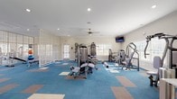 Located at 93 Dune Lakes Circle in Santa Rosa Beach, Florida, this single-floor fitness center is a comprehensively outfitted facility designed to support a wide range of workout routines. The space is organized into distinct functional zones, seamlessly connected across an open, generously proportioned floor plan finished with a vibrant blue and earth-toned modular carpet throughout.

The cardio section features a row of treadmills, elliptical trainers, and a stationary bike, all positioned along a wall of large windows that flood the area with natural light and offer views of the surrounding community. Full-length mirrors along one wall enhance the sense of spaciousness and allow users to monitor their form during exercise.

The strength training area is well-appointed with a comprehensive dumbbell rack, multiple cable and selectorized resistance machines, and a dedicated stretching and functional fitness zone equipped with stability balls and a weight bench. Ornate white lattice partition screens add an architectural design element that defines zones while maintaining visual openness throughout the space.

Recessed lighting and ceiling fans ensure the environment remains bright and comfortable. Wall-mounted televisions are positioned for visibility from both the cardio and strength areas. Beach-themed artwork adorns the walls, reinforcing the coastal character of the Santa Rosa Beach location.