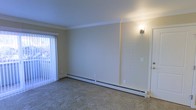 This single-floor residence offers a clean, functional layout designed for comfortable everyday living. The main living area is finished with wall-to-wall carpeting that adds warmth underfoot, while crown molding along the ceiling lends a subtle touch of architectural detail. Neutral, light-toned walls create a bright and welcoming ambiance throughout the space.

A prominent feature of the living area is the large window dressed with vertical blinds, allowing for generous natural light and views to the outdoors. The room is equipped with baseboard heating units running along the base of the walls, ensuring efficient climate control throughout the colder months. A wall-mounted sconce light fixture provides supplementary ambient lighting, adding a practical yet understated design element.

The overall layout is open and uncluttered, offering versatility for a variety of furniture arrangements and uses. The single-floor plan ensures ease of navigation throughout the space, making it accessible and straightforward. The consistent use of neutral tones across the walls, ceiling, and flooring contributes to a cohesive and calm interior atmosphere, well-suited for those seeking a simple, move-in-ready environment.