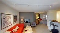 This single-floor studio is thoughtfully designed to maximize its open-plan layout, seamlessly blending living and sleeping areas into one cohesive space. The living area anchors the room with a vibrant orange sofa paired with a light-toned coffee table, creating an inviting focal point perfect for relaxing or entertaining. A flat-screen television rests atop a sleek media console nearby, completing the lounge setup. The sleeping area transitions naturally from the living space, featuring a comfortable bed accented with bold orange pillows and a warm wooden dresser that adds a touch of natural texture. A decorative mosaic-style mirror serves as a striking accent piece above the dresser, adding visual depth and personality to the sleeping nook. A plush neutral carpet runs throughout, softening the space and contributing to its overall warmth. The walls are finished in a sophisticated gray palette, complementing the contemporary furnishings while providing a calm, neutral backdrop. Track lighting installed along the ceiling provides flexible, well-distributed illumination across the entire space. The textured ceiling adds subtle character, and the cohesive design ensures every corner of the studio feels purposeful and polished.