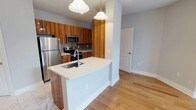 This single-floor space showcases a thoughtfully designed open-concept layout that seamlessly connects the kitchen and living areas. The kitchen is a standout feature, outfitted with warm, rich wood cabinetry that adds depth and character to the space. Stainless steel appliances — including a full-size refrigerator, microwave, and range — provide a polished, contemporary look complemented by a subway tile backsplash. A large white quartz peninsula anchors the kitchen, offering ample prep space and an integrated sink, making it both functional and visually striking. Pendant lighting with globe-shaped fixtures hangs above the peninsula, casting a warm, inviting glow. The transition from tile flooring in the kitchen to light-toned hardwood floors in the adjacent living area creates a natural visual flow throughout the space. Neutral gray walls keep the atmosphere bright and versatile, while the clean architectural lines and open sightlines contribute to an airy, modern ambiance. A paneled interior door leads to additional areas of the unit, hinting at a well-organized and functional floor plan. Overall, the space balances contemporary design with warm, natural materials for a refined and livable environment.