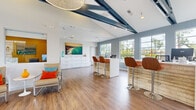 Located at 1456 Serfas Club Dr in Corona, California, the Sierra Del Oro Apartments leasing office is a single-story space designed to make a strong first impression. The interior is defined by soaring vaulted ceilings accented with bold, exposed navy steel beams that add an industrial-modern character to the otherwise light and airy environment. Warm hardwood-style flooring runs throughout, complementing the crisp white walls and soft blue-gray accent tones.

Upon entry, guests are welcomed by a spacious foyer featuring the Sierra Del Oro branded entryway mat and oversized glass double doors that invite natural light deep into the space. A stylish pendant light fixture anchors the entry area, while large multi-pane windows along the perimeter frame views of the outdoor surroundings and fill the interior with abundant daylight.

The main area serves as both a welcoming lounge and a functional workspace. A casual seating arrangement with upholstered chairs and vibrant orange and teal accent pillows provides a comfortable area for guests. Adjacent to this is a textured wood-panel bar counter with modern swivel stools, functioning as an informal consultation station. A white reception desk with workstations is centrally positioned, and a dedicated refreshment nook with counter seating, a mini-fridge, and beverage station adds a hospitality touch. Coastal-themed artwork and curated décor complete the inviting, resort-inspired ambiance.