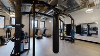 Located at 215 N First Ave E in downtown Duluth, Minnesota, this fitness center occupies a single floor and offers a comprehensive exercise environment within a characterful, industrial-style setting. The space is defined by its exposed stone and brick walls, open-beam ceilings with visible ductwork and piping, and pendant lighting that reinforces the raw, urban aesthetic. Rubber flooring throughout provides a durable, functional surface suited for a wide range of workouts.

The exercise room is thoughtfully divided into distinct training zones. A cardio section houses multiple treadmills equipped with interactive touchscreen displays, along with a stationary spin bike, an elliptical, and a rowing machine — all positioned near windows that bring in natural light and offer views of the surrounding downtown Duluth streetscape. A strength training area features a full dumbbell rack with an adjustable weight bench and framed wall art that adds a polished touch to the utilitarian environment.

A central multi-functional training rig serves as the focal point of the space, complete with a heavy punching bag, TRX suspension straps, battle ropes, and a pull-up station. A large wall-mounted mirror enhances the sense of openness. The overall ambiance is energetic and purposeful, blending historic architectural character with modern fitness amenities.