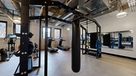 Located at 215 N First Ave E in downtown Duluth, Minnesota, this fitness center occupies a single floor and offers a comprehensive exercise environment within a characterful, industrial-style setting. The space is defined by its exposed stone and brick walls, open-beam ceilings with visible ductwork and piping, and pendant lighting that reinforces the raw, urban aesthetic. Rubber flooring throughout provides a durable, functional surface suited for a wide range of workouts.

The exercise room is thoughtfully divided into distinct training zones. A cardio section houses multiple treadmills equipped with interactive touchscreen displays, along with a stationary spin bike, an elliptical, and a rowing machine — all positioned near windows that bring in natural light and offer views of the surrounding downtown Duluth streetscape. A strength training area features a full dumbbell rack with an adjustable weight bench and framed wall art that adds a polished touch to the utilitarian environment.

A central multi-functional training rig serves as the focal point of the space, complete with a heavy punching bag, TRX suspension straps, battle ropes, and a pull-up station. A large wall-mounted mirror enhances the sense of openness. The overall ambiance is energetic and purposeful, blending historic architectural character with modern fitness amenities.