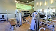 The Racquet Club's fitness center is a thoughtfully designed workout facility that combines functionality with a clean, welcoming aesthetic. The main floor is home to a generous array of strength training equipment, including a selection of cable and resistance machines as well as a dedicated free weight area with a full dumbbell rack. The flooring is a durable, textured surface suited for high-traffic athletic use, and recessed ceiling lights ensure the space is consistently and evenly illuminated throughout. Wall-mounted sconces add a refined touch to the overall ambiance. A standout architectural feature is the open mezzanine level, enclosed by sleek glass panel railings that allow natural sightlines between floors and contribute to the facility's bright, airy feel. A staircase connects the two levels, and the upper level mirrors the layout below with additional equipment visible through the glass. Built-in shelving along the walls provides convenient storage for personal items or accessories. The space is further enhanced by large mirrors that visually expand the room and assist with exercise form. Overall, the fitness center at The Racquet Club delivers a professional and well-organized environment suited for a comprehensive workout experience.