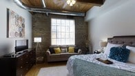 This single-floor space showcases an industrial-loft aesthetic, combining raw architectural elements with thoughtfully curated furnishings. The bedroom is defined by its striking exposed brick walls, which lend texture and historic character to the room, complemented by a warm wood-planked ceiling supported by visible beams. Metallic ductwork runs along the ceiling, reinforcing the space's industrial charm while adding an authentic urban edge.

A large, multi-pane window serves as the focal point of the room, flooding the space with natural light and offering views of the surrounding urban environment. Horizontal blinds provide adjustable privacy without sacrificing brightness.

The bedroom is comfortably furnished with a generously sized bed dressed in layered neutral bedding, accompanied by a small loveseat positioned near the window, decorated with yellow and grey accent pillows. Warm hardwood flooring anchors the space, while a soft area rug adds comfort underfoot. A tripod floor lamp and ceiling light fixture with multiple bulbs provide ambient lighting options. Dark wood dressers offer practical storage, completing the balanced mix of industrial and livable elements throughout this distinctive space.
