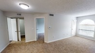 This single-floor unit offers a straightforward, functional layout designed for comfortable everyday living. The main living area is finished with warm, neutral-toned carpet and painted in crisp white, creating a clean and open feel. A wall-mounted thermostat and ceiling-mounted light fixture are present, along with ceiling vents for climate control. A textured popcorn ceiling runs throughout the carpeted spaces, adding a classic residential character. An interior doorway connects the living space to an additional carpeted room, which features sliding panel doors that allow for natural light and access to an outdoor area.

The kitchen is situated adjacent to the entry and features white cabinetry, white countertops, and light tile flooring. Black appliances, including a refrigerator and electric range, provide contrast against the bright cabinetry. A peninsula counter separates the kitchen from the main living space, offering a casual dining or prep area. A single-basin sink with a window above completes the kitchen setup.

The bathroom includes a full tub and shower combination with white tile surround, gold-toned fixtures, and a coordinating towel ring. A dedicated laundry closet houses a stacked washer and dryer unit set on hardwood-style flooring, adding practical convenience to the space.