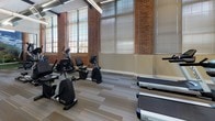 This fitness center seamlessly blends industrial character with modern amenities, creating an inviting and functional workout environment. The main exercise area is defined by striking exposed brick walls and tall, large-pane windows fitted with adjustable blinds, allowing abundant natural light to fill the space throughout the day. Overhead, exposed ductwork and linear pendant lighting complement the industrial aesthetic, while striped, neutral-toned carpet tiles cover the floors throughout.

The cardio zone is generously outfitted with a variety of equipment, including treadmills, ellipticals, upright and recumbent stationary bikes, and additional cross-training machines arranged in orderly rows. Wall-mounted televisions provide entertainment during workouts. Full-length glass walls partition sections of the fitness center, maintaining an open, airy feel while defining distinct workout zones.

A standout feature is the enclosed putting green and golf simulator area, enclosed by floor-to-ceiling glass panels. This dedicated space features artificial turf and a large scenic golf course mural, offering a unique recreational option within the fitness facility. Additional open floor space accommodates strength and stretching equipment, rounding out the comprehensive offerings of this well-appointed fitness center.