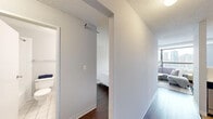 This thoughtfully laid-out urban unit offers an efficient and comfortable single-floor living experience. A bright, narrow entry hallway leads into the main living space, where large floor-to-ceiling windows frame sweeping city skyline views, flooding the room with natural light. The open-concept living area features rich hardwood flooring, soft neutral tones, and a seamless transition between the sitting area and sleeping space, maximizing the sense of openness throughout.

The kitchen is functionally designed with white cabinetry, white countertops, stainless steel appliances — including a refrigerator, electric range, and over-the-range microwave — and warm tile flooring, offering a clean and practical workspace. A textured ceiling adds subtle character throughout the unit.

The well-appointed closet provides generous built-in storage with multiple hanging rods across two sections and a central shelving unit with drawers, making excellent use of available space. Dark hardwood flooring continues through this area, maintaining design consistency.

The bathroom is clean and bright, featuring a white vanity with a large mirror, a tiled shower-tub combination, and crisp white finishes throughout. Overall, this unit presents a cohesive, modern aesthetic with practical amenities and remarkable urban views.