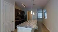 This two-floor space showcases a contemporary open-concept layout that seamlessly connects the kitchen and living areas. The kitchen is a standout feature, centered around a generous island topped with striking granite countertops displaying bold veining patterns in neutral tones. Three elegant cylindrical pendant lights hang above the island, providing focused task lighting while adding a decorative touch. Dark espresso cabinetry lines the kitchen walls, creating a sharp contrast against the lighter countertop surfaces and walls. Stainless steel appliances, including a refrigerator, microwave, and range, lend a sleek, modern aesthetic. A track lighting system overhead offers additional illumination throughout the space. The kitchen flows naturally into the living area, where hardwood-style flooring extends underfoot and a ceiling fan is visible near a window fitted with blinds, allowing natural light to filter in. The overall color palette of soft grays, warm beiges, and dark wood tones creates a balanced, modern ambiance. The two-floor layout provides additional living space beyond what is immediately visible, suggesting a well-organized and functional floor plan throughout the home.