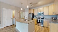 Located at 9101 Stony Point Pkwy in Richmond, Virginia, this single-floor residence offers a well-designed layout with a seamless flow between its main living areas. The kitchen serves as a central highlight, featuring a large granite-topped island with an integrated sink, pendant lighting, and a full suite of stainless steel appliances including a refrigerator, electric range, and over-the-range microwave. Light wood-tone cabinetry extends to the ceiling, providing generous storage, while wood-look flooring adds warmth throughout the space. The open layout connects the kitchen directly to the living area, which is bright and airy, benefiting from natural light streaming through glass panel doors and a ceiling fan overhead. The bedrooms are carpeted for comfort, each offering a neutral color palette and access to walk-in closets with built-in shelving. The bathroom includes a full soaking tub with subway tile surround, wood-look flooring, and a granite-topped vanity that mirrors the finishes found throughout the unit. A dedicated laundry area is conveniently accessible from the kitchen. Overall, the space presents a clean, contemporary aesthetic with consistent finishes and a functional, open design.
