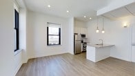 This 1-bedroom, 1-bathroom unit is designed with a clean, modern aesthetic and an open-concept floor plan that seamlessly connects the living and kitchen areas. The living space features light-toned hardwood flooring and crisp white walls, creating a bright and airy atmosphere enhanced by recessed lighting overhead. A well-positioned window frames views of the surrounding cityscape, allowing natural light to fill the room.

The kitchen is thoughtfully appointed with white cabinetry extending to the ceiling, stainless steel appliances including a refrigerator, stove, and built-in microwave, and a peninsula countertop with a granite-style surface that doubles as a breakfast bar. A stainless steel sink is integrated into the peninsula, and pendant lighting above adds both function and style.

The overall design emphasizes clean lines and neutral tones, offering a versatile canvas suited to a variety of interior styles. The open layout maximizes the sense of space, making the unit feel expansive and well-connected. The combination of modern finishes and practical design elements makes this a well-appointed urban living space.