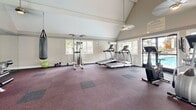 Located at 2611-2635 Yorba Linda Blvd in Fullerton, CA, this fitness center offers a well-appointed exercise space designed to support a comprehensive workout routine. The room is defined by striking vaulted ceilings with exposed beam detailing, which lend an open, airy feel and contribute to the spacious atmosphere throughout. Carpet tile flooring in muted tones covers the exercise area, providing comfort and traction underfoot.

Natural light flows generously through multiple large windows and glass sliding doors positioned along multiple walls, offering pleasant views of an outdoor pool and surrounding landscaping. This abundance of natural light, combined with the high ceilings, creates a bright and welcoming environment.

The fitness center is equipped with a diverse selection of exercise equipment, including treadmills, elliptical trainers, a stationary bike, a cable and weight machine, a bench press station, and a heavy punching bag with a speed bag station. A wall-mounted flat-screen television provides entertainment during workouts, while a full-length mirror along one wall enhances the sense of space and allows for form monitoring. A wall-mounted air conditioning unit ensures a comfortable environment year-round. Fitness center rules, workout calendars, and informational postings are displayed on the walls, adding a functional and organized touch to the space.