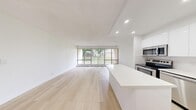 This well-appointed 3-bedroom, 1-bathroom unit is thoughtfully designed across a single floor, offering a clean, modern aesthetic throughout. The open-concept kitchen and living room form the heart of the space, anchored by light hardwood-style flooring that runs continuously through the unit. The kitchen is outfitted with sleek white cabinetry, white tile backsplash, stainless steel appliances including a range, microwave, and refrigerator, and a central island with a white countertop that doubles as additional prep and dining space. Recessed lighting illuminates the kitchen and living area, enhancing the bright, airy atmosphere. A wall of large sliding floor-to-ceiling windows in the living area floods the space with natural light and provides unobstructed views of a lush green lawn and mature trees. The three bedrooms are bright and well-proportioned, with the primary bedroom offering generous closet space featuring mirrored sliding doors. Each bedroom benefits from natural light via large windows. The full bathroom is finished in crisp white tile with a tub-shower combination. A dedicated laundry area and hallway complete the functional layout, making this a well-rounded and move-in ready space.