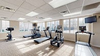 Located at 1420 Key Pkwy in Frederick, Maryland, this fitness center offers a dedicated exercise room designed to support a full range of workout routines. The space is finished with a drop ceiling fitted with recessed panel lighting that provides consistent, even illumination throughout. Large multi-pane windows line the walls on multiple sides, flooding the room with natural light and offering views of the surrounding outdoor environment, creating an open and energizing atmosphere. The flooring consists of neutral-toned carpet, lending warmth and comfort underfoot. The cardio section is well-appointed with multiple True-brand treadmills and elliptical trainers, each equipped with individual digital consoles, as well as upright and recumbent stationary bikes. The strength training area features cable and pulley machine systems alongside weight benches, offering users a comprehensive resistance workout. Wall-mounted flat-screen televisions are positioned at multiple angles for easy viewing during exercise. A wall mirror enhances the sense of space, while a hand sanitizer station and restroom access contribute to a clean and functional environment. A glass-paneled door leads to an adjoining area, further expanding accessibility within the facility.