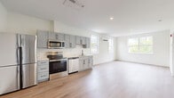 Located at 3 Railroad Street in Newmarket, NH, this thoughtfully designed one-bedroom, one-bathroom unit offers a clean, modern aesthetic across its well-appointed single floor. The open-concept kitchen and living area serves as the heart of the space, featuring light hardwood flooring, crisp white walls, and abundant natural light from multiple windows. The kitchen is outfitted with gray shaker-style cabinetry, black hardware accents, a full suite of stainless steel appliances including a refrigerator, electric range, over-the-range microwave, and dishwasher, as well as a white countertop and undermount sink. A wall-mounted mini-split unit provides efficient climate control in the living area, which also enjoys views of the surrounding neighborhood. The bedroom is carpeted for comfort, features a window overlooking green trees, and connects conveniently to the full bathroom. The bathroom is finished with gray cabinetry, a white countertop vanity, black hardware fixtures, light hardwood flooring, and a full soaking tub. A hallway connects the main living spaces, adding to the functional flow of the layout. The unit presents a fresh, move-in-ready interior with consistent modern finishes throughout.