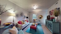 This thoughtfully designed single-floor residence offers a welcoming and vibrant living experience across several well-defined spaces. The living room serves as the heart of the home, featuring plush carpeting, a textured ceiling, and an abundance of natural light from a window overlooking greenery. Eclectic décor — including circular wall art, a bold teal area rug, a vivid red ottoman, and colorful throw pillows — creates an energetic yet comfortable atmosphere. A dedicated media area with a stylish teal credenza and wall art adds functionality and character to the room.

The kitchen is efficiently laid out with white cabinetry, laminate countertops, tile flooring, and a full suite of appliances including a dishwasher, electric range, and built-in microwave. Decorative touches above the cabinets add a personalized feel.

Two bedrooms provide restful retreats, each carpeted and accented with tasteful artwork. The primary bedroom features a ceiling fan, a walk-in closet, and warm, layered bedding. The second bedroom benefits from natural light through a window with blinds.

The bathroom includes a vanity with white cabinetry, a large mirror with overhead lighting, tile flooring, and patterned wallcovering that adds a distinctive design element to the space.
