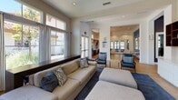 Located at 25550 River Bend Dr in Yorba Linda, California, this single-story community clubhouse offers a well-appointed gathering space designed for comfort and socialization. The open-concept lounge area serves as the centerpiece, featuring elevated ceilings with recessed lighting and a stylish chandelier overhead. Generous windows dressed with striped floor-to-ceiling drapes line the walls, flooding the interior with natural light and providing views of the surrounding outdoor areas, including a community pool. The lounge is furnished with a spacious neutral-toned sectional sofa, coordinating accent chairs in blue and beige tones, and large ottomans arranged atop a navy area rug over wood-look flooring. Adjacent to the main lounge, intimate dining nooks offer round wood tables surrounded by patterned accent chairs, framed by expansive windows overlooking the pool and landscaped grounds. French doors provide seamless access to the outdoor spaces. A kitchenette or service area is visible toward the rear of the space, adding functional utility. The overall ambiance is contemporary and inviting, with a cohesive blue and neutral color palette, ample seating arrangements, and an abundance of natural light that creates a bright, welcoming atmosphere throughout.