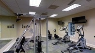 This fitness center offers a practical and well-organized workout space designed to accommodate a range of exercise needs. The room features a drop-panel ceiling with recessed fluorescent lighting that ensures even, bright illumination throughout the space. The walls are finished in a neutral warm beige tone, contributing to a clean and focused environment.

The facility is equipped with a multi-station cable weight machine, providing users with versatile strength training options. Cardio equipment includes an elliptical trainer, a stationary bike, and additional cardio machines positioned near the perimeter of the room. A wall-mounted flat-screen television is positioned at eye level, allowing users to stay entertained during workouts.

The flooring consists of a durable patterned carpet tile, suitable for a high-traffic exercise environment. A mirrored wall panel along one side of the room enhances the sense of space and allows users to monitor their form during exercise. A window provides a connection to the exterior, allowing natural light to supplement the overhead fixtures. Overall, the space is thoughtfully arranged to maximize functionality and comfort for its users.