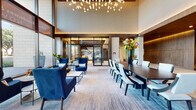This elegantly designed lobby and amenity space exudes a polished, upscale ambiance throughout. The grand main lobby commands attention with soaring double-height ceilings and floor-to-ceiling glass walls that flood the interior with natural light and offer open views to the surrounding streetscape. A sculptural chandelier of cascading globe pendants serves as a striking focal point overhead, complementing the refined mix of wood-paneled accents, light stone tile flooring, and plush area rugs. The lounge area is furnished with rich navy blue velvet seating arranged around sleek coffee tables, while a large communal dining table with tailored upholstered chairs provides a sophisticated gathering space. A dedicated mailroom features warm-toned wood millwork, coffered ceilings adorned with globe-cluster chandeliers, and an expansive bank of resident mailboxes alongside a compact seating arrangement. The reception area presents a dramatic stone-clad desk set against wood-paneled walls and curated artwork. A gallery-style corridor lined with framed artwork and rich wood paneling leads to private leasing offices, where bold abstract murals and pendant lighting create an inspiring, design-forward environment. Every detail reflects a commitment to luxurious, contemporary design.