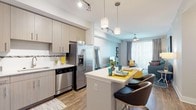 This thoughtfully designed single-floor residence offers a seamless blend of modern style and functional living. The open-concept layout connects the kitchen and living area, creating a bright and cohesive space ideal for both everyday living and entertaining. The kitchen is equipped with stainless steel appliances, light wood-tone cabinetry, a tile backsplash, quartz countertops, and a peninsula island with bar seating, illuminated by pendant lighting overhead. Track lighting adds a contemporary touch to the ceiling. Wood-style flooring flows throughout the main living areas, anchoring the warm, modern aesthetic. The living room features a ceiling fan and ample space for a full furniture arrangement, with access to a glass-paneled sliding door that invites natural light. The two bedrooms are generously sized, each with ceiling fans, wood-style floors, and large windows. One bedroom includes a walk-in closet and direct access to an en suite bathroom. The bathrooms are clean and modern — one featuring a soaking tub with a glass partition and a floating vanity, and the other offering a walk-in shower with recessed lighting and a light stone-toned vanity. The overall ambiance is contemporary and well-lit, with a neutral color palette accented by warm wood tones.