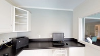 This single-floor office space is thoughtfully designed for productivity, featuring a built-in L-shaped workstation with a sleek dark countertop that wraps along two walls, providing generous desk space for computer equipment and peripherals. White lower cabinetry beneath the countertop offers integrated storage, while open upper shelving on one wall adds additional organizational capacity.

The walls are painted in a calming light blue-gray tone, complemented by crisp white crown molding and trim throughout, lending the space a clean, professional aesthetic. Dark woven-texture flooring anchors the room and contrasts neatly with the white cabinetry and light walls.

A distinctive pass-through window connects this office to an adjacent common area, enhancing communication and visual openness between spaces. A white-paned French door beside the pass-through provides a formal entry point while maintaining transparency into the neighboring room. Natural light filters in through window treatments, contributing to a bright, airy atmosphere. A framed piece of colorful artwork on the wall adds a subtle decorative touch, balancing the functional nature of the space with a welcoming ambiance.